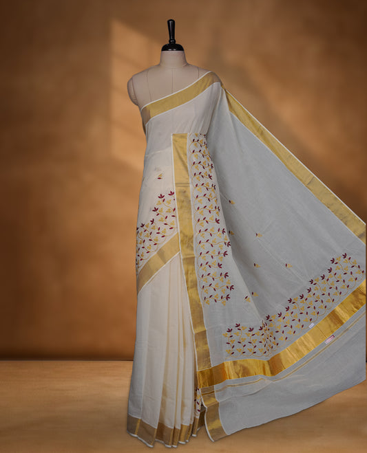 Off-white colour cotton Kerala saree featuring a classic gold kasavu border, highlighted with delicate maroon and mustard floral vine embroidery along the pallu and borders, and paired with a matching blouse.
