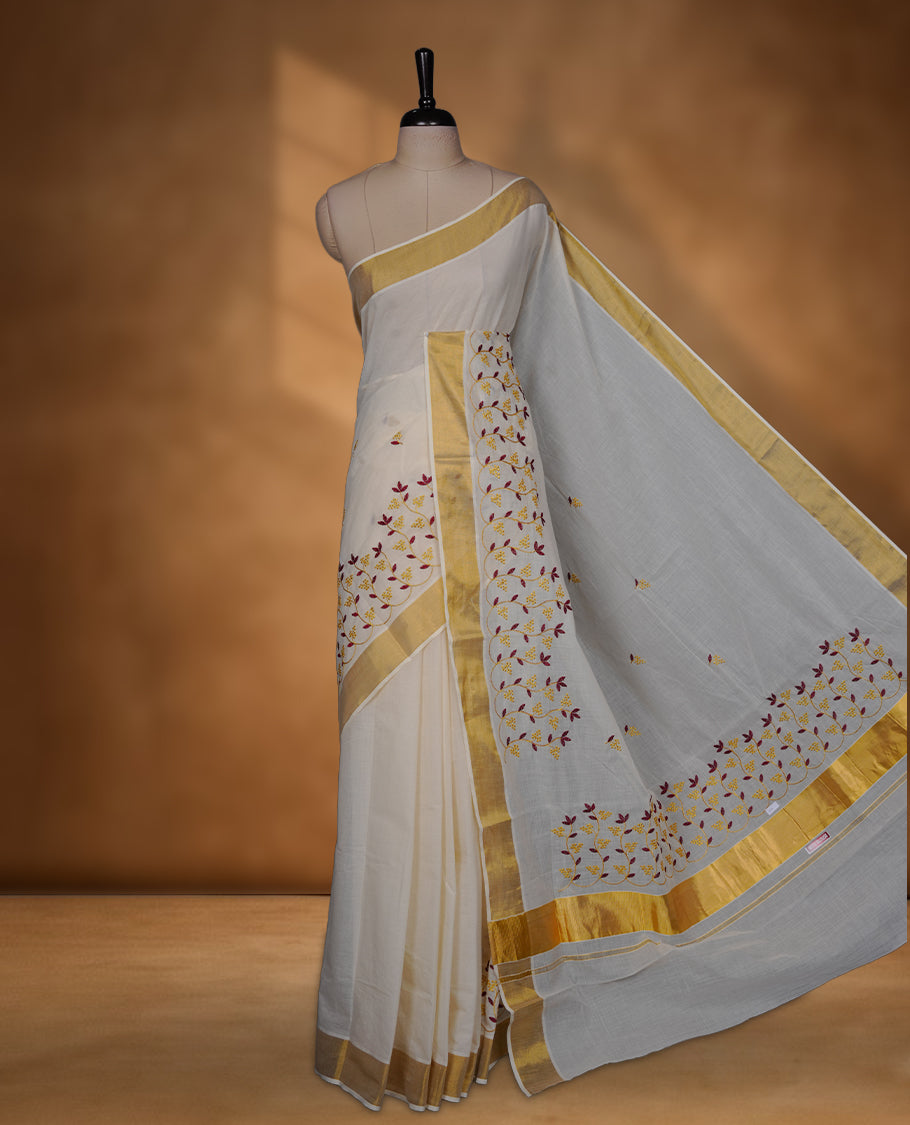 Off-white colour cotton Kerala saree featuring a classic gold kasavu border, highlighted with delicate maroon and mustard floral vine embroidery along the pallu and borders, and paired with a matching blouse.