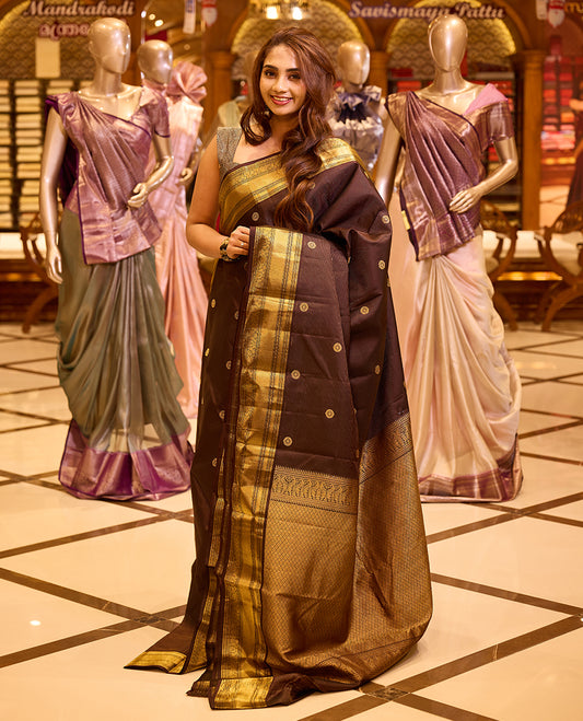 Graceful deep chocolate brown colour Kancheepuram silk saree adorned with delicate gold zari motifs across the body, beautifully finished with a broad traditional gold zari border, exuding timeless elegance and paired with a matching blouse piece