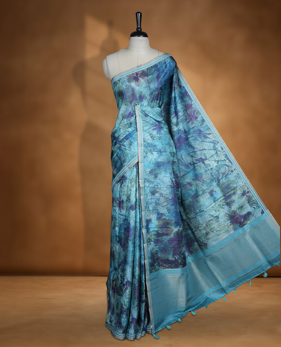 Teal blue colour semi tussar silk saree featuring an abstract floral printed body with a rich self border and tasselled pallu, paired with a matching blouse.