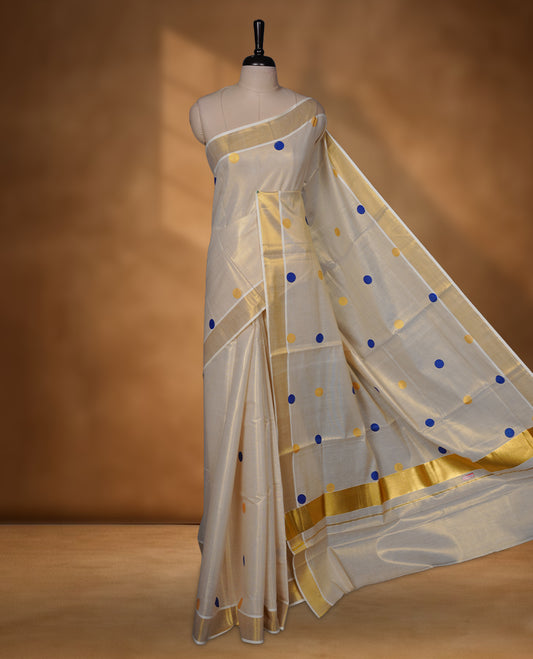 Off-white colour Tissue Kerala saree featuring playful blue and gold polka buttas across the body, finished with a classic gold kasavu border and paired with a matching blouse.