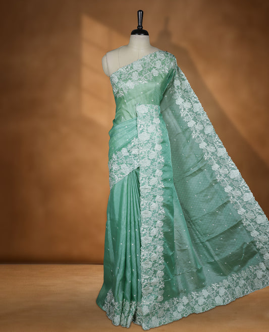 Light Green colour organza designer saree featuring a subtle self patterned body and an intricately scalloped floral thread embroidered border all around, complemented by a coordinating embroidered pallu and paired with a matching blouse.