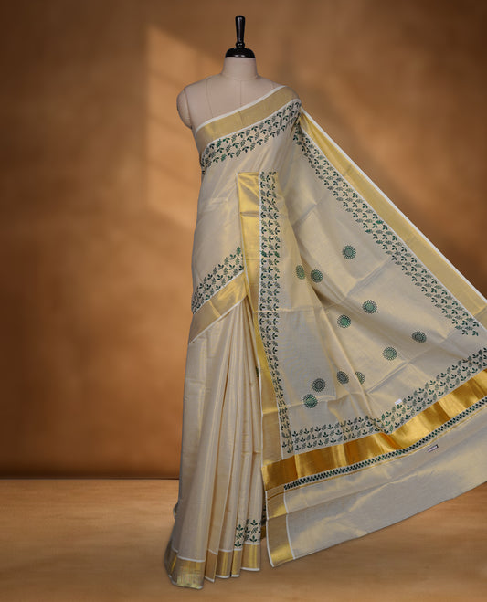 Off-white colour tissue Kerala saree featuring a classic gold kasavu border, highlighted with elegant green leafy and floral motifs across the body and pallu, and paired with a matching blouse.