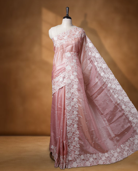 Pastel peach colour organza designer saree featuring a subtle self patterned body and an intricately scalloped floral thread embroidered border all around, complemented by a coordinating embroidered pallu and paired with a matching blouse.