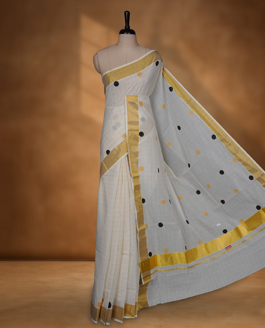 Off-white colour cotton Kerala saree with a classic gold kasavu border, highlighted by subtle gold checks and playful black & mustard polka dot motifs across the body and pallu, paired with a matching blouse.