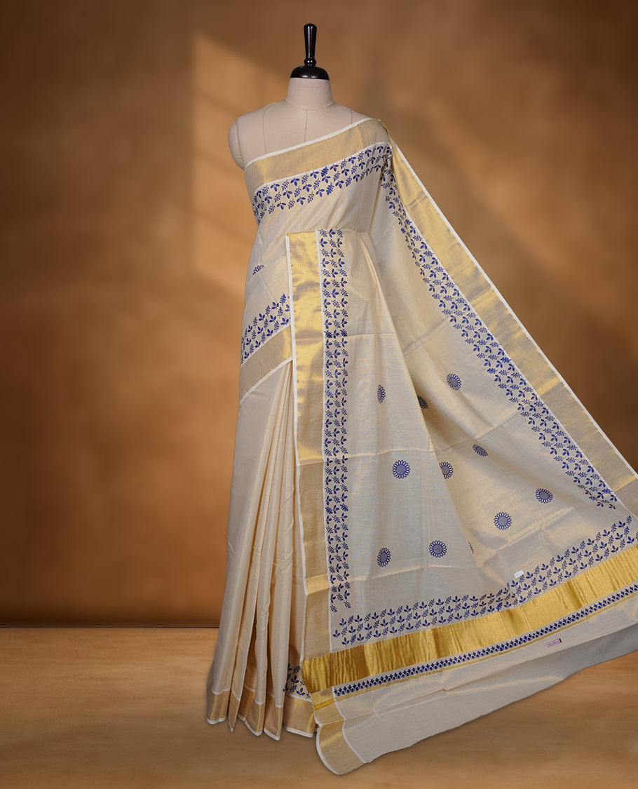 Off-white colour tissue Kerala saree with a rich gold kasavu border, adorned with delicate royal blue floral motifs across the body and pallu, paired with a matching blouse.