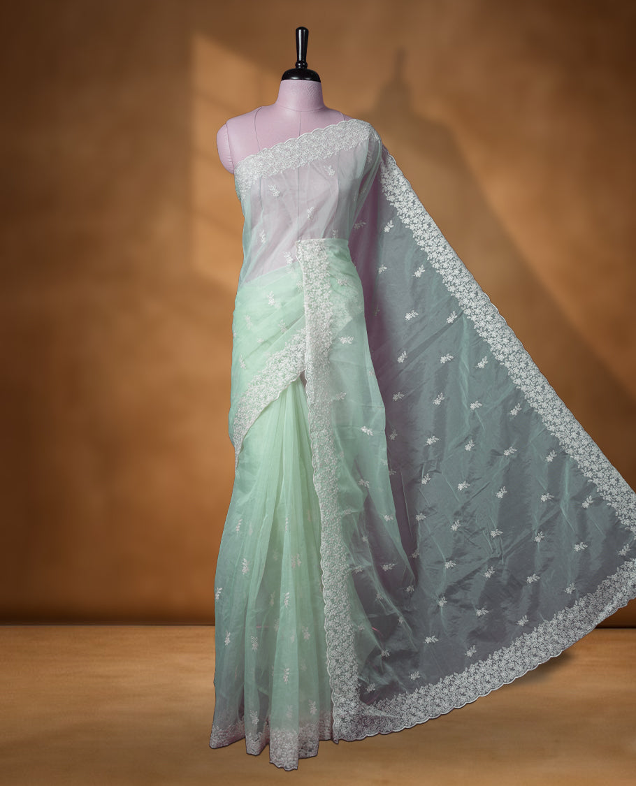 Pastel green colour organza designer saree featuring a subtle self patterned body and an intricately scalloped floral thread embroidered border all around, complemented by a coordinating embroidered pallu and paired with a matching blouse.