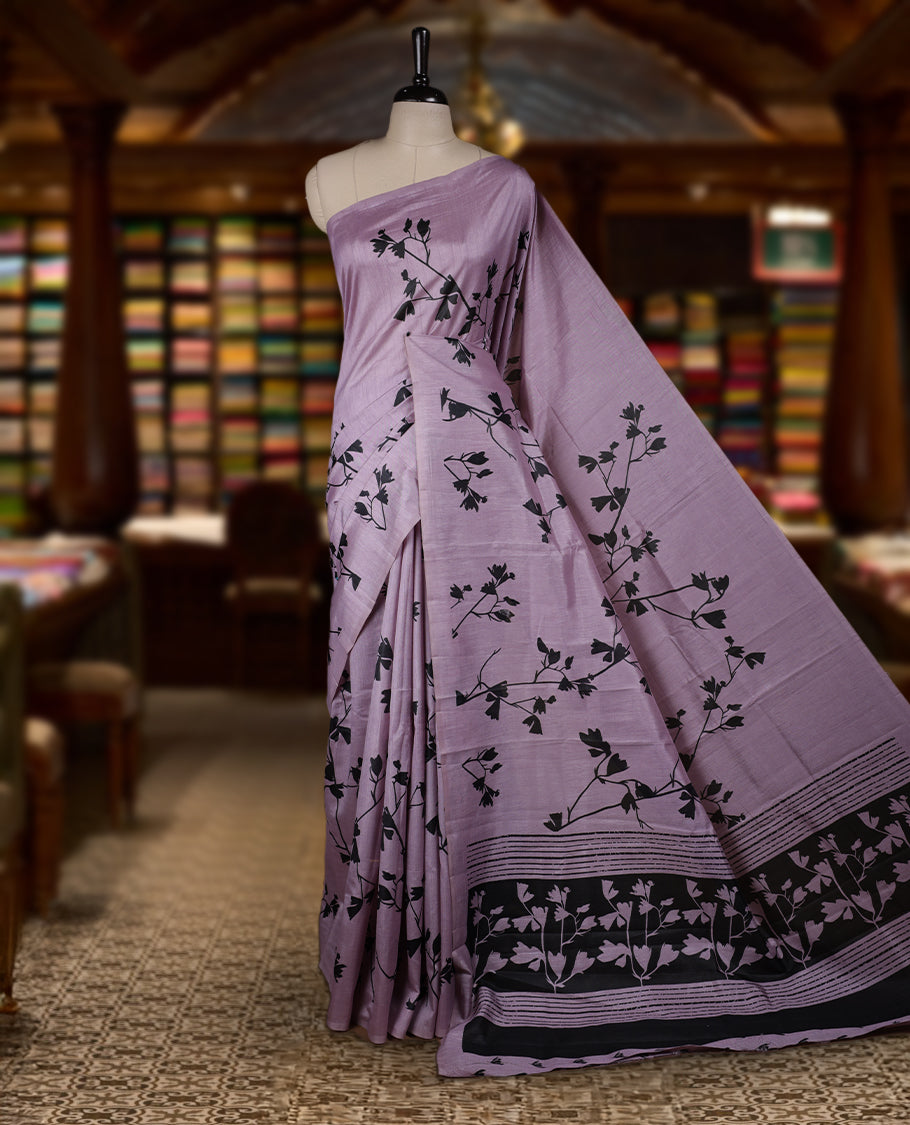 Pastel Lavender colour Semi tussar silk saree adorned with black floral prints on the body and an elegant pallu & Comes with a matching blouse piece for a classy look.
