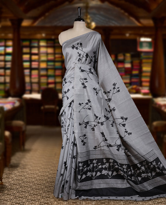 Pastel Grey colour Semi tussar silk saree adorned with black floral prints on the body and an elegant pallu & Comes with a matching blouse piece for a classy look.
