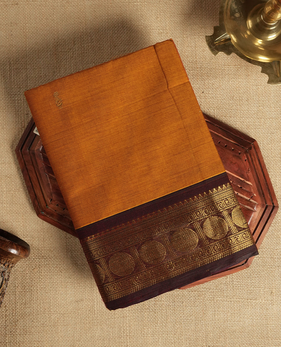 Mustard Orange colour Chettinad cotton saree adorned with small woven motifs on the body, The saree features a deep maroon border highlighted with intricate golden zari circular patterns and comes with a matching blouse piece.