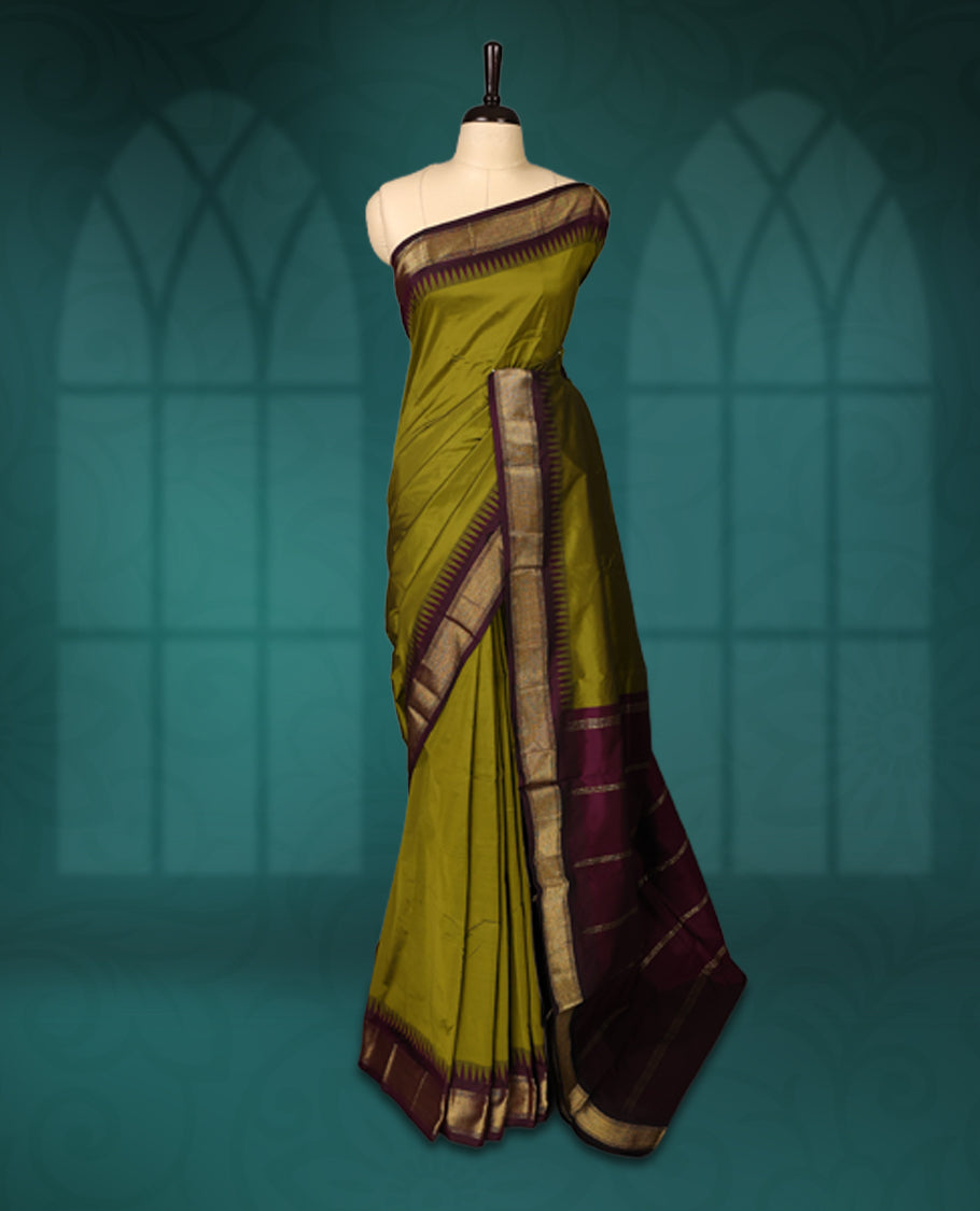 Olive green semi silk saree with plain body, vibrant deep purple pallu and matching purple border adorned with golden zari, accented with temple motifs along the edges, Paired with a matching blouse, perfect for festive occasions.
