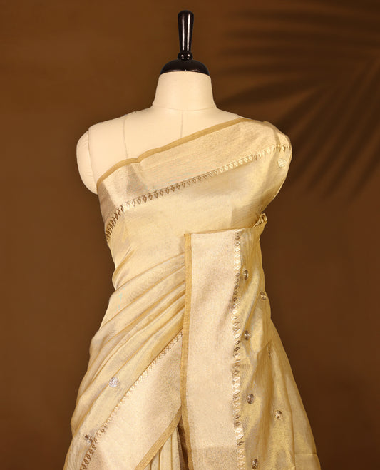 Light gold colour Organza Kerala saree featuring a luminous body with a subtle golden sheen, enhanced by a sleek zari border and scattered sequin like floral motifs, Paired elegantly with a matching blouse piece.