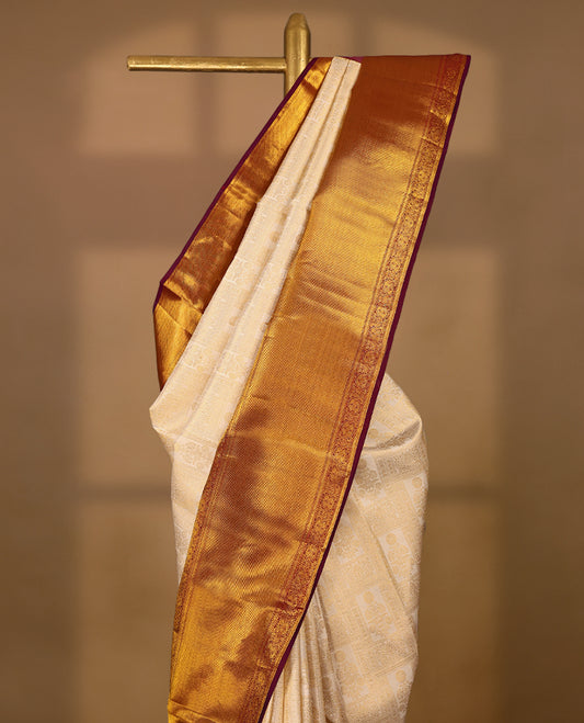 Off-white Colour traditional Kancheepuram silk saree adorned with antique zari floral motifs arranged in a symmetric grid on the body, featuring a contrasting border with floral, peacock, and chakram motifs, and paired with a matching blouse.