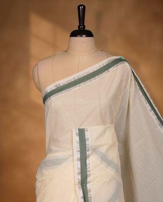 Off-white colour cotton Kerala saree featuring a subtle green border with silver zari accents, a clean plain body and pallu, and paired with a matching blouse.