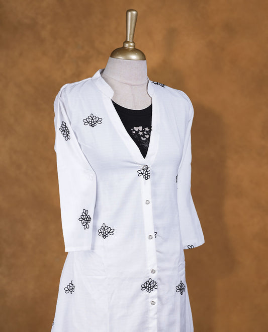 White embroidered shirt dress with band collar, contrast black inner layer at neckline, front button placket, three-quarter sleeves, and intricate black border embroidery at hem