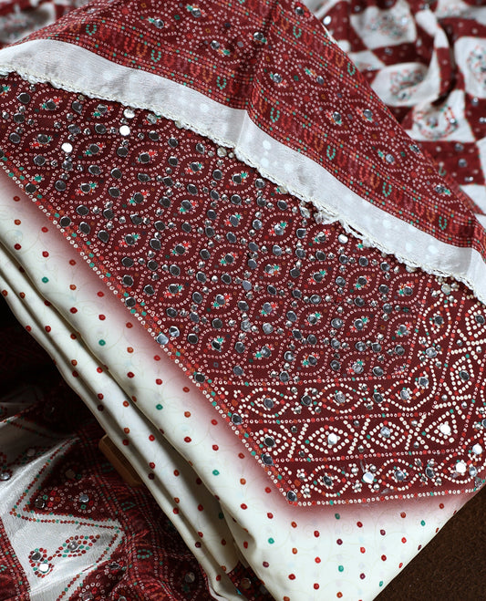 Off white colour churidhar material adorned with mirror work and maroon colour traditional bandhani patterns, paired with a green and maroon colour dotted off white top, plain bottom, and an elegant matching dupatta.