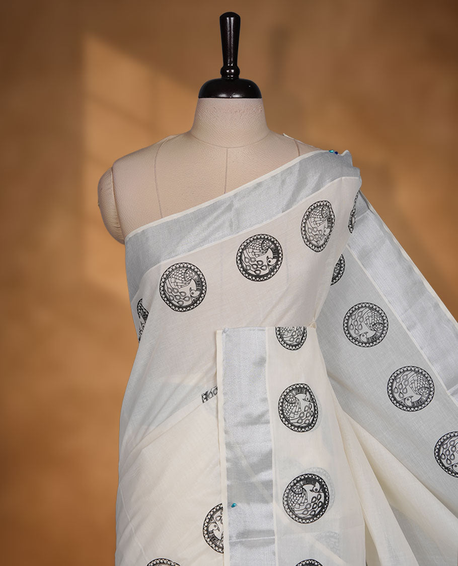 Off-white colour cotton Kerala saree featuring a subtle silver kasavu border, adorned with classic black cirlcle peacock motifs across the body and pallu, and paired with a matching blouse.