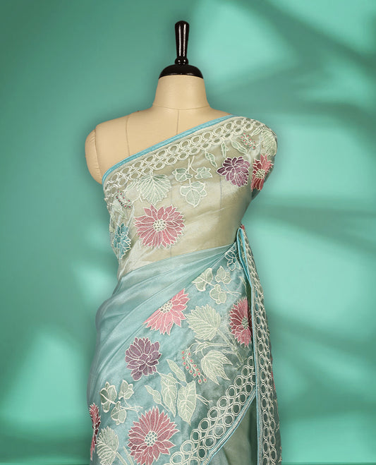 Aqua Blue colour organza designer saree featuring delicate pastel floral embroidery in pink, mint green, and white across the body, with a scalloped border accentuated by floral threadwork, and a gracefully embroidered pallu paired with matching blouse.