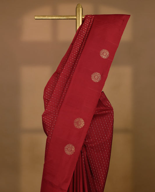 Deep red colour traditional Kancheepuram silk saree adorned with vertical rows of small golden motifs on the body, featuring a self-coloured simple border with floral chakra motifs, and paired with a matching blouse.