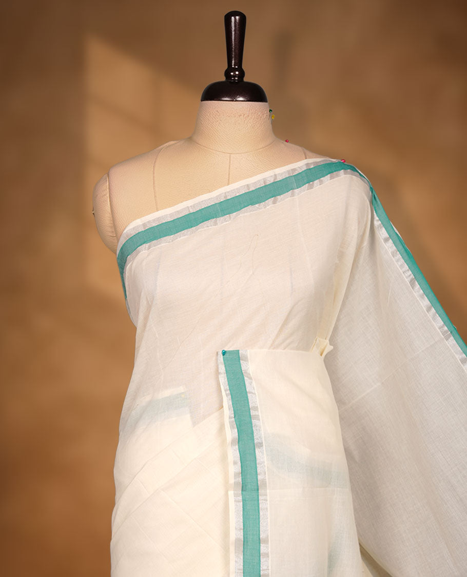 Off-white colour cotton Kerala saree featuring a crisp white body with a refreshing green kasavu border, detailed with subtle silver accents along the edges, and paired with a matching blouse.