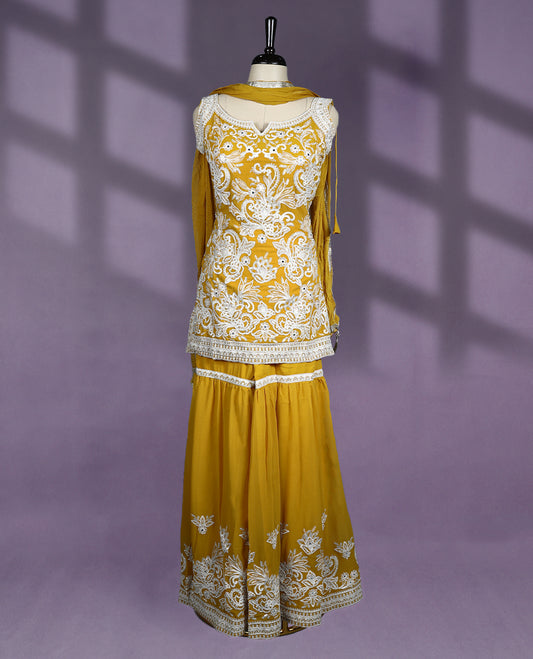 Mustard colour sharara set features a beautifully embellished sweetheart neckline with cutout detail and sleeveless design, adorned with intricate white embroidery throughout the kurti and hem of the flared sharara pants, paired with a matching dupatta
