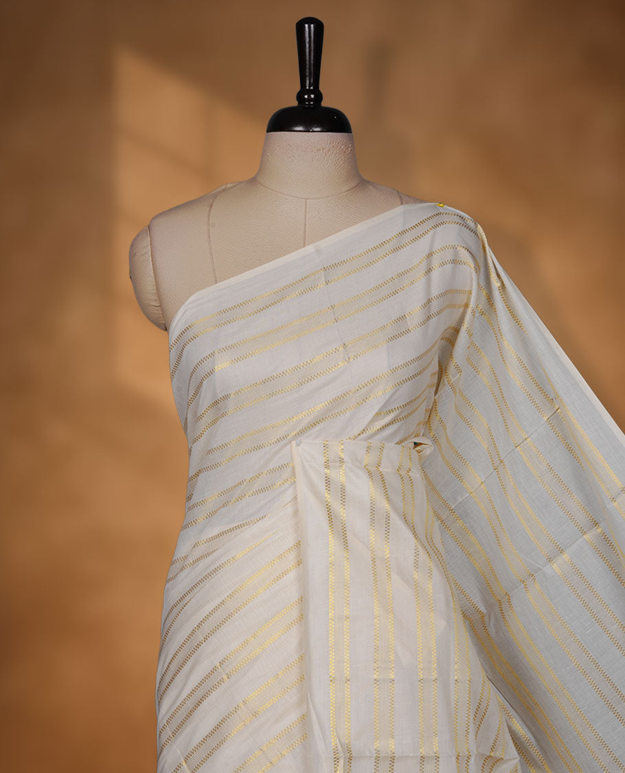 Off-white colour cotton Kerala saree featuring subtle gold zari stripe detailing across the body, finished with elegant tassels on the pallu, and paired with a matching blouse.