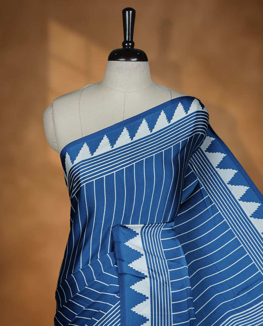 Blue colour semi modal saree highlighted with white triangle border accents and sleek striped detailing across the drape, featuring a bold oversized white circular motif on the pallu, paired with a matching printed blouse.