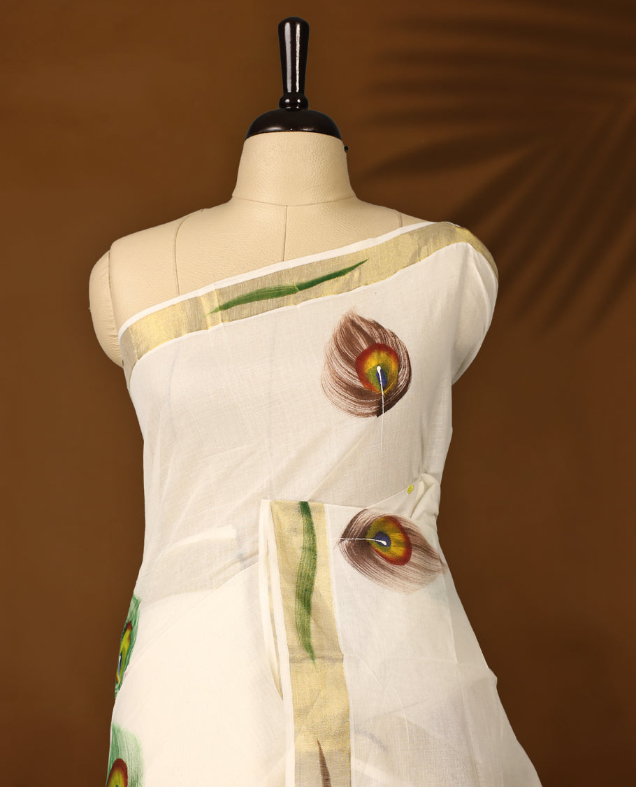 Elegant off white colour cotton Kerala saree with hand-painted peacock feathers and scenic pallu artwork, highlighted by a golden zari border and pallu, paired with a matching blouse for a graceful festive look.