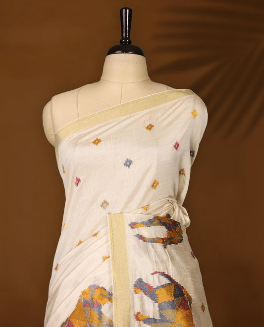 Cream colour Tussar saree adorned with multicolour geometric elephant motifs and delicate thread buttas across the body, enhanced with a subtle gold zari border, paired with a matching blouse.
