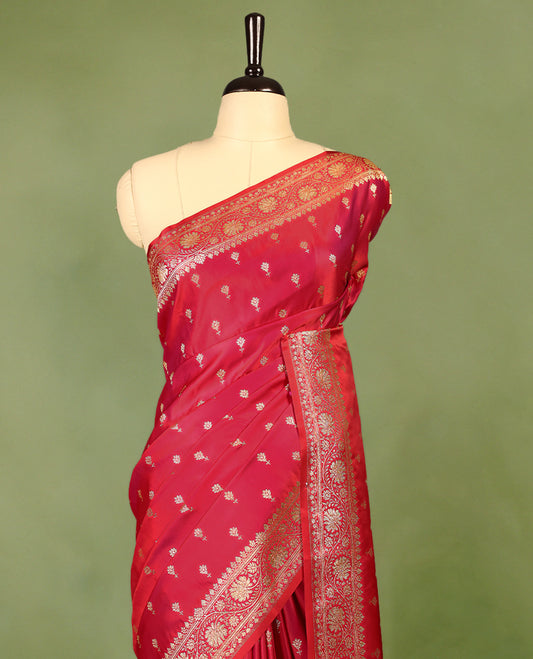 Pinkish red colour semi Banaras satin crepe saree featuring gold zari woven flower motifs across the body, complemented by an intricately designed traditional banarasi border and richly woven pallu, paired with a matching blouse piece.