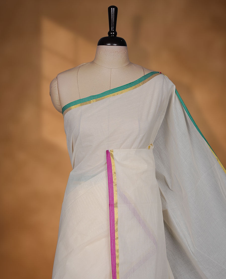 Off-white colour cotton Kerala saree, featuring a classic gold kasavu border highlighted with subtle green and magenta accents, finished with a soft, airy drape and paired with a matching blouse.