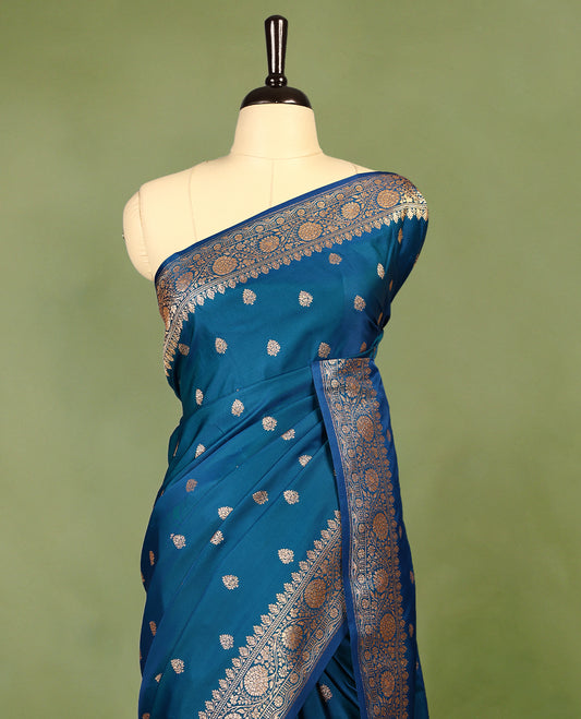 Peacock blue colour semi Banaras satin crepe saree featuring gold zari woven floral motifs across the body, complemented by an intricately designed traditional banarasi border and richly woven pallu, paired with a matching blouse piece.