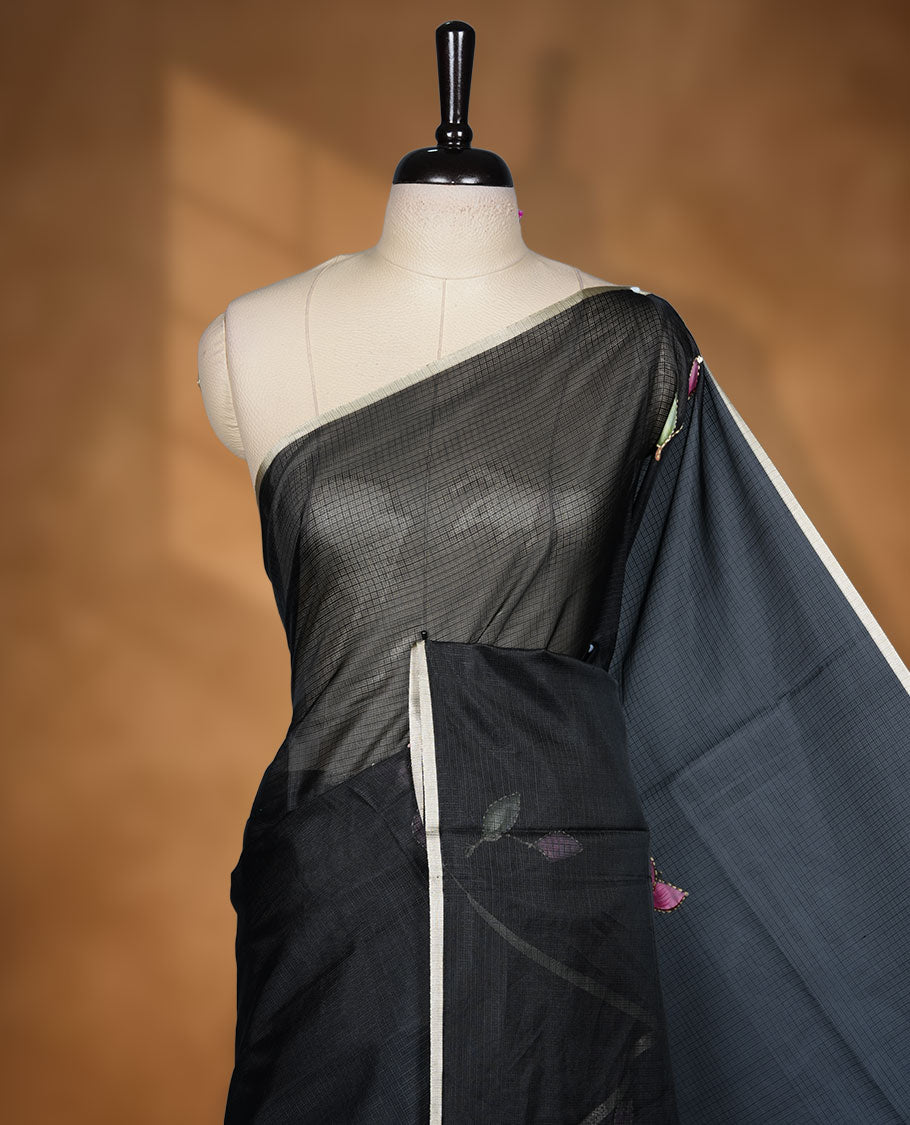 Black colour semi tusser silk saree adorned with fine floral embroidery spread across the body, highlighted by subtle contrast accents along the border, paired with a matching blouse.