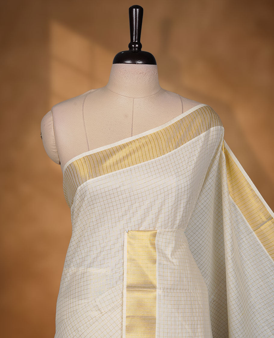 Off-white colour cotton Kerala saree featuring a subtle gold checkered weave, highlighted with a rich traditional gold kasavu border along the pallu and pleats, and paired with a matching blouse.