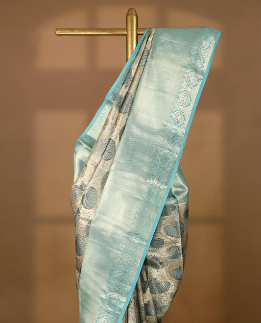 Silver Colour Kancheepuram tissue silk saree featuring intricate silver and blue zari leaf motifs on the body, gracefully complemented by a light blue traditional peacock and floral design border, and paired with a matching blouse.