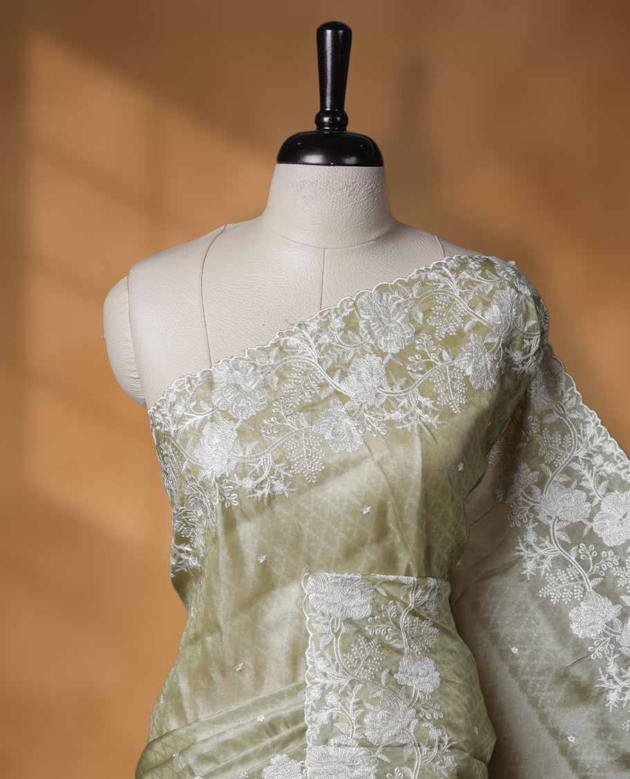 Pastel olive colour organza designer saree featuring a subtle self patterned body and an intricately scalloped floral thread embroidered border all around, complemented by a coordinating embroidered pallu and paired with a matching blouse.