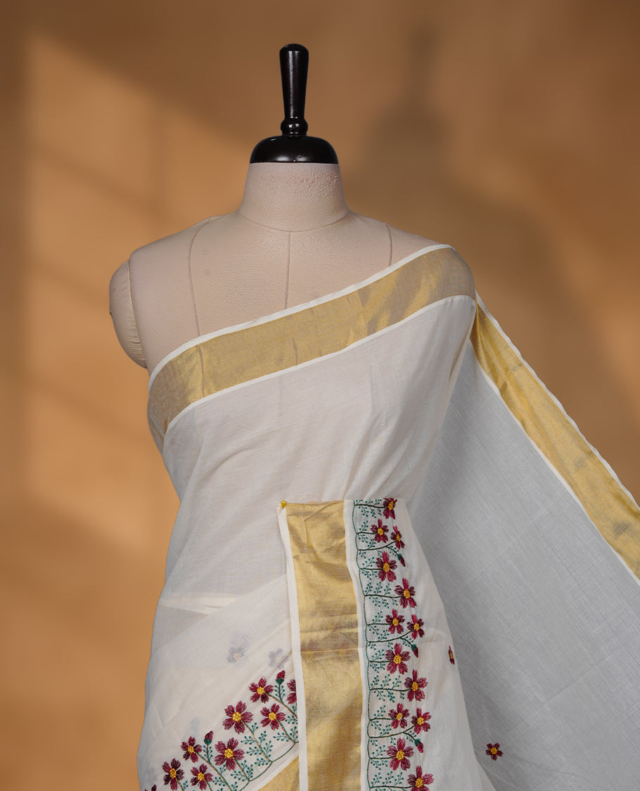 Off-white colour Kerala saree with a classic gold kasavu border, delicately embroidered floral motifs in red and green across the body and pallu, finished with a subtle golden sheen for timeless elegance.