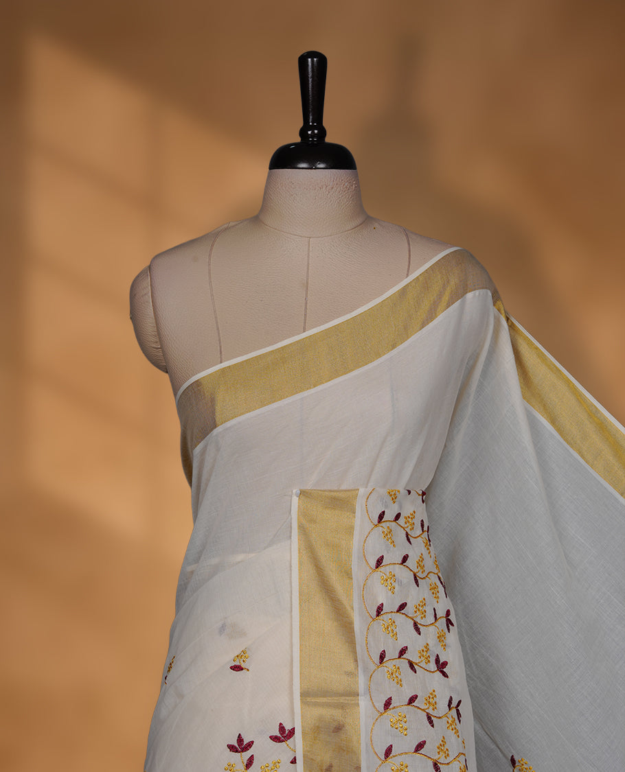 Off-white colour cotton Kerala saree featuring a classic gold kasavu border, highlighted with delicate maroon and mustard floral vine embroidery along the pallu and borders, and paired with a matching blouse.
