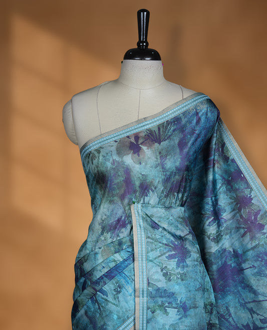 Teal blue colour semi tussar silk saree featuring an abstract floral printed body with a rich self border and tasselled pallu, paired with a matching blouse.