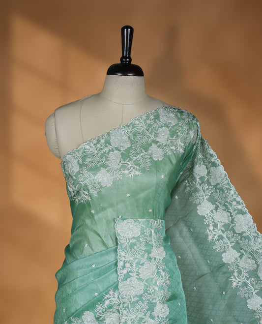 Light Green colour organza designer saree featuring a subtle self patterned body and an intricately scalloped floral thread embroidered border all around, complemented by a coordinating embroidered pallu and paired with a matching blouse.