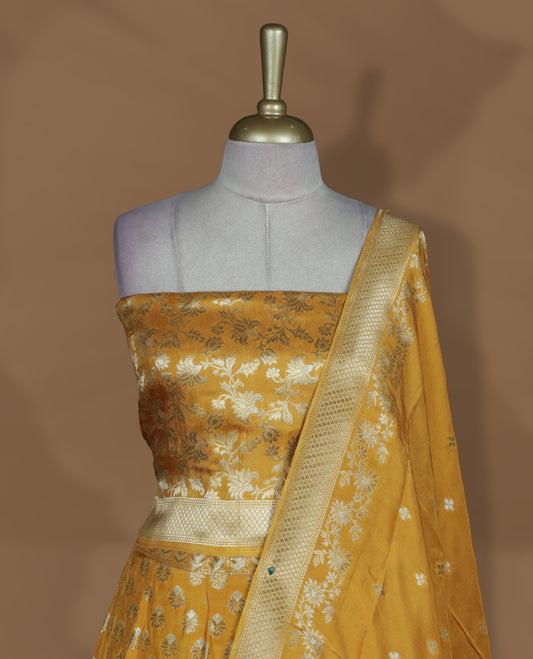 Yellow colour Banarasi lehenga set with a stitched flared lehenga featuring all‑over zari butti motifs and rich woven border, paired with an unstitched floral zari blouse piece and matching dupatta with zari border and tasselled edges.