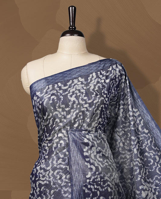Greyish blue colour semi jute tissue saree featuring white floral prints across the body, adorned with intricate geometric woven borders and a graceful floral work pallu & Comes with a matching blouse piece.