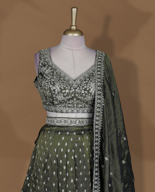Olive green and black ombre effect lehenga set with stitched sleeveless V‑neck heavily embroidered blouse, matching stitched flared lehenga featuring intricate zari and sequin borders, and coordinated dupatta with embellished edge.
