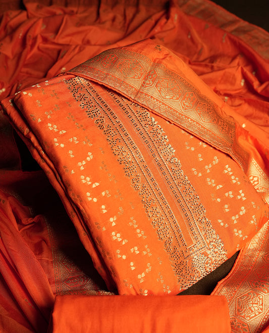 Orange colour churidar material adorned with small golden stone work on the neckline, featuring intricate zari floral motifs on the body, rich zari woven border and dupatta, paired with matching plain bottom.