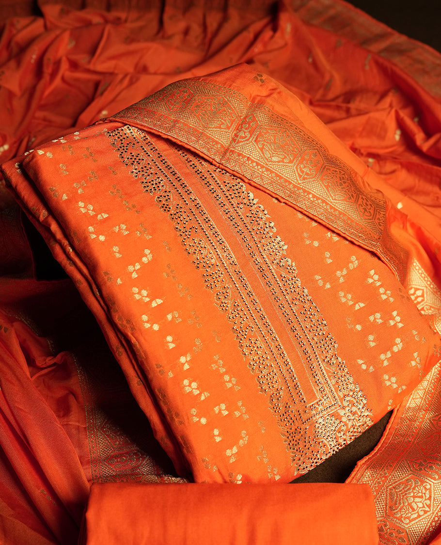 Orange colour churidar material adorned with small golden stone work on the neckline, featuring intricate zari floral motifs on the body, rich zari woven border and dupatta, paired with matching plain bottom.
