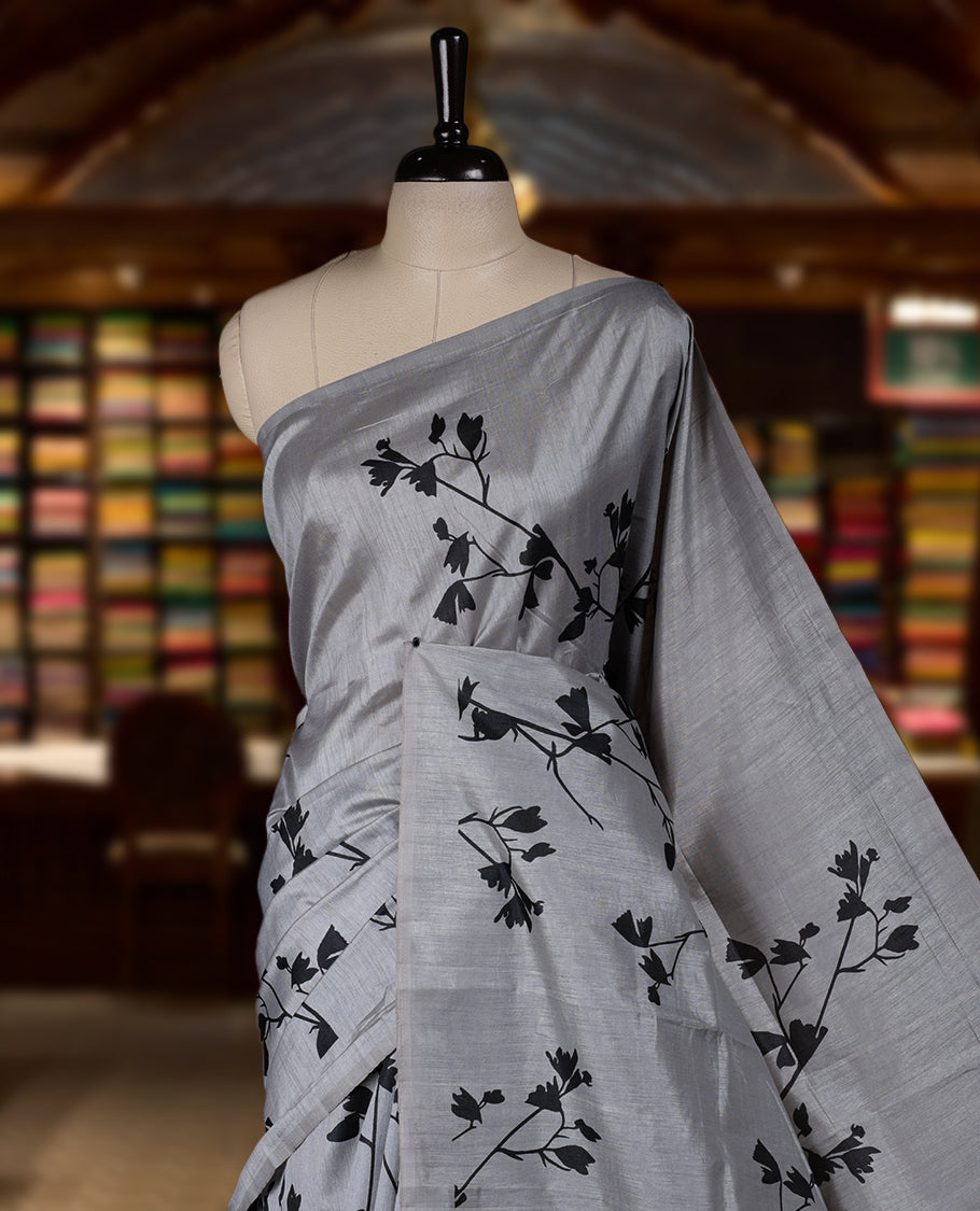 Pastel Grey colour Semi tussar silk saree adorned with black floral prints on the body and an elegant pallu & Comes with a matching blouse piece for a classy look.