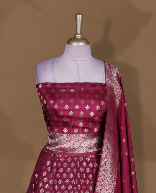 Wine colour Banarasi lehenga set with a stitched flared lehenga featuring all‑over zari butti motifs and rich woven border, paired with an unstitched floral zari blouse piece and matching dupatta with zari border and tasselled edges.
