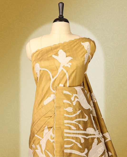 Golden Brown colour semi tussar saree adorned with elegant white applique work in leaf and floral designs across the body and border, paired with a matching blouse.