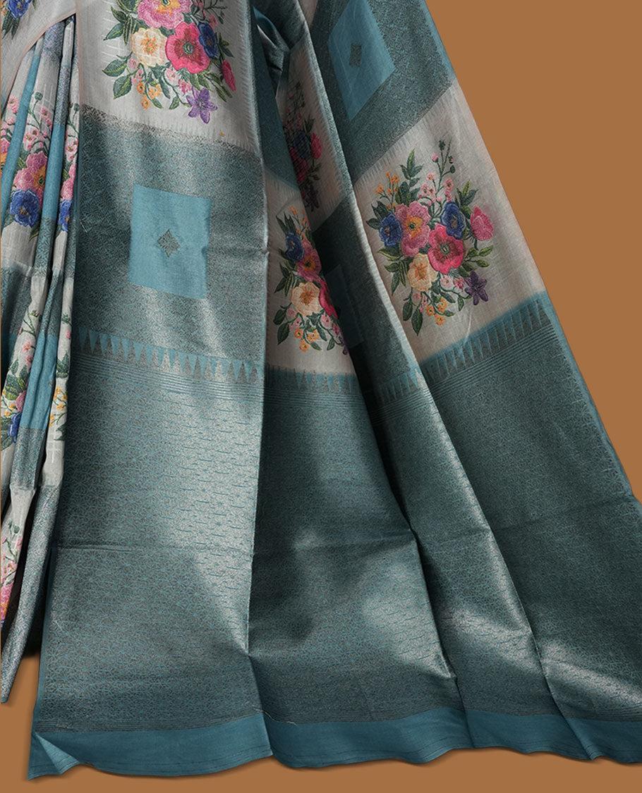 Blue color semi tussar Banarasi saree featuring vibrant multi-color floral prints across the body, with geometric zari patterns, and a matching blouse detailed with silver woven buttas.