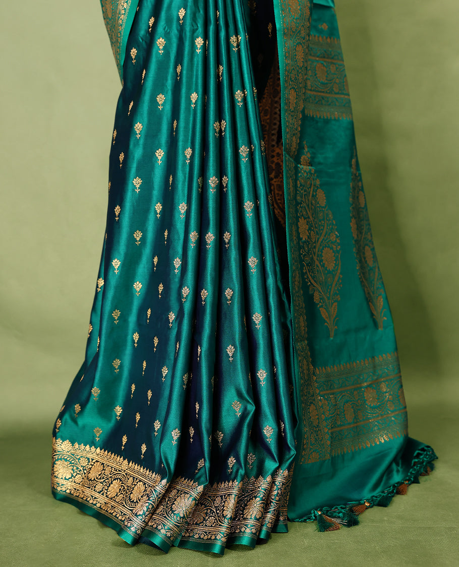 Deep sea green colour semi Banaras satin crepe saree featuring gold zari woven flower motifs across the body, complemented by an intricately designed traditional banarasi border and richly woven pallu, paired with a matching blouse piece.